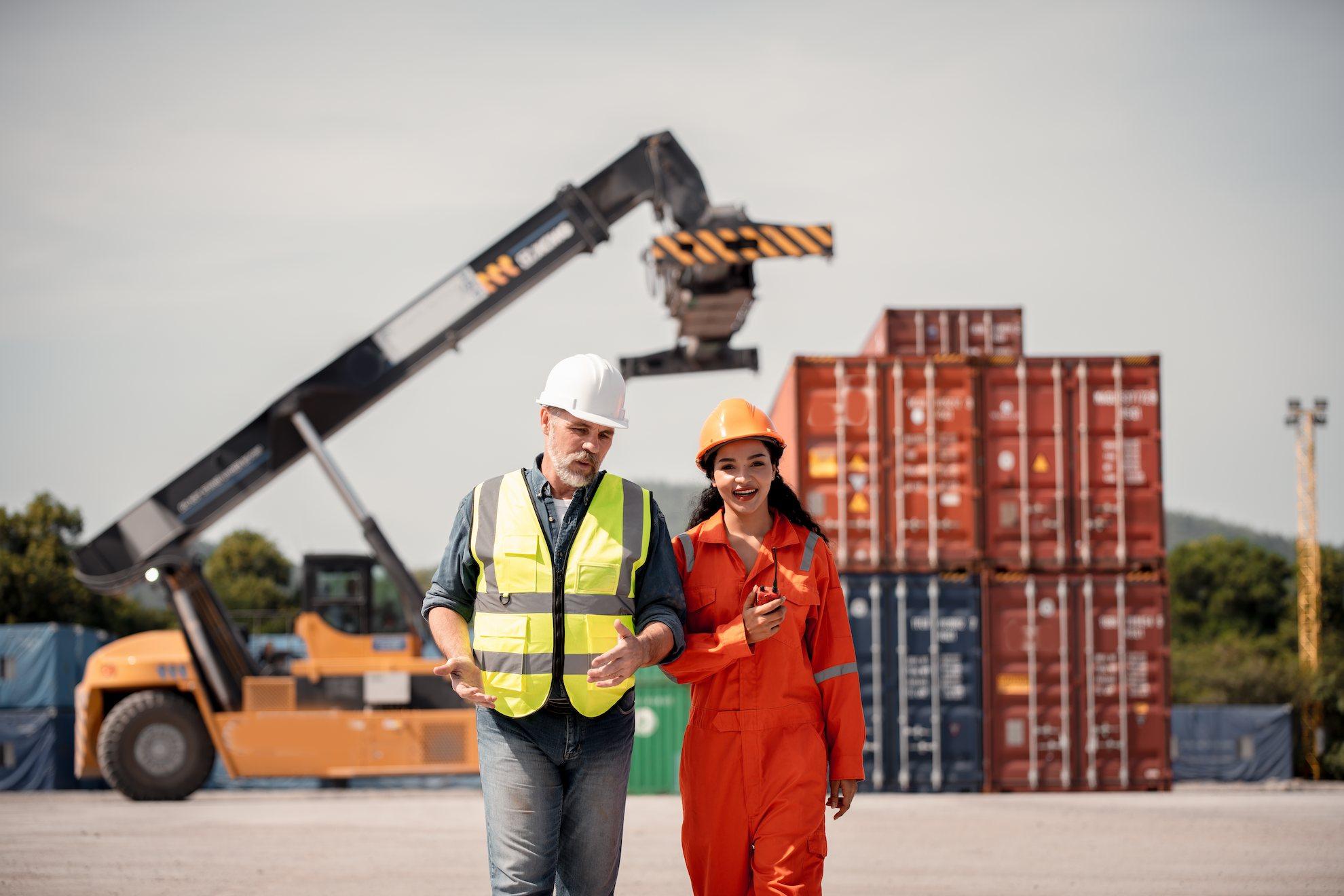 Two workers at a Dubai shipping yard discuss logistics operations in front of cargo containers and a crane, illustrating industrial trade license Dubai activities.